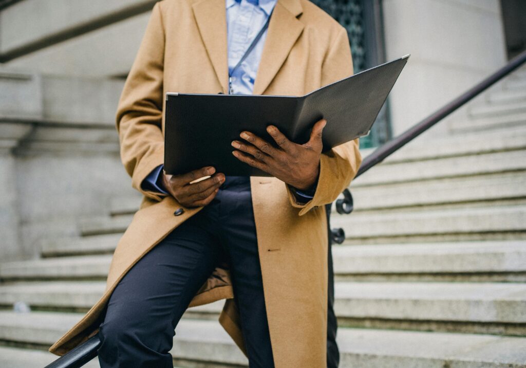 A well-dressed man in a tan coat reads a document on city steps, embodying professionalism.