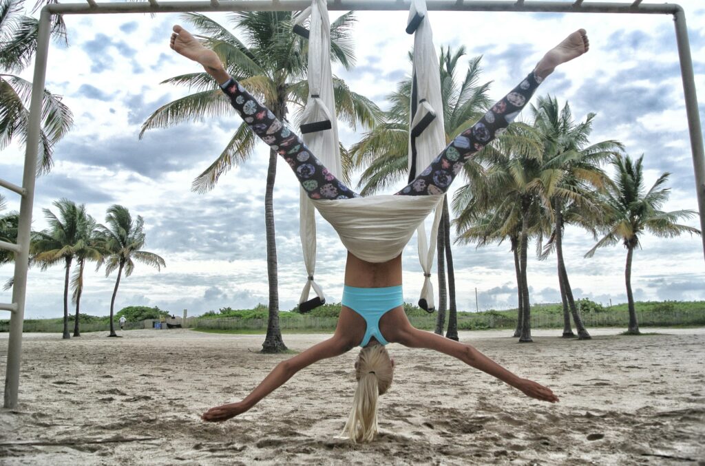 Blonde woman performing aerial yoga on a sandy beach surrounded by palm trees.