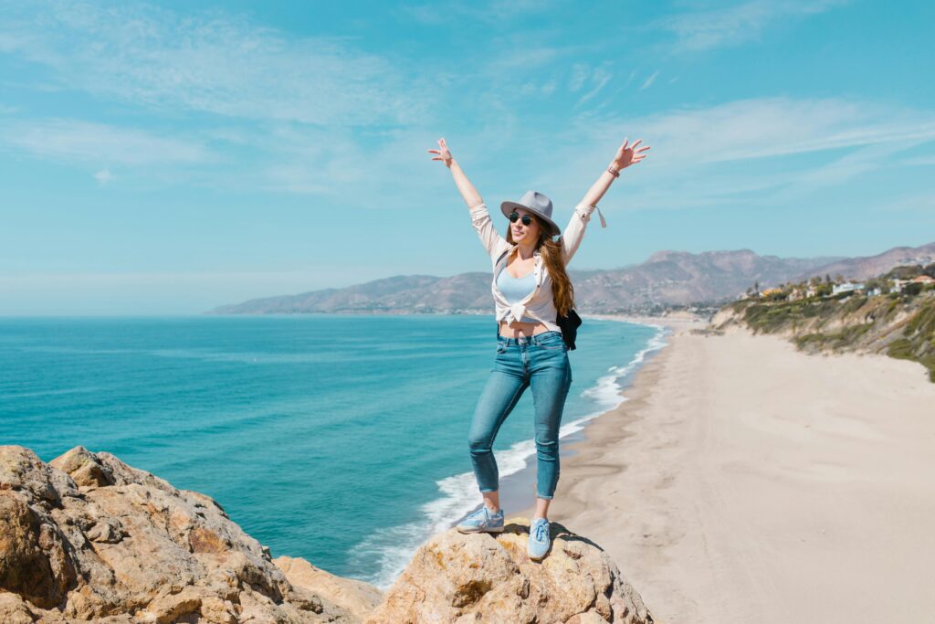 Smiling woman poses on a rocky cliff overlooking a sunny beach and ocean, embracing the coastal beauty.
