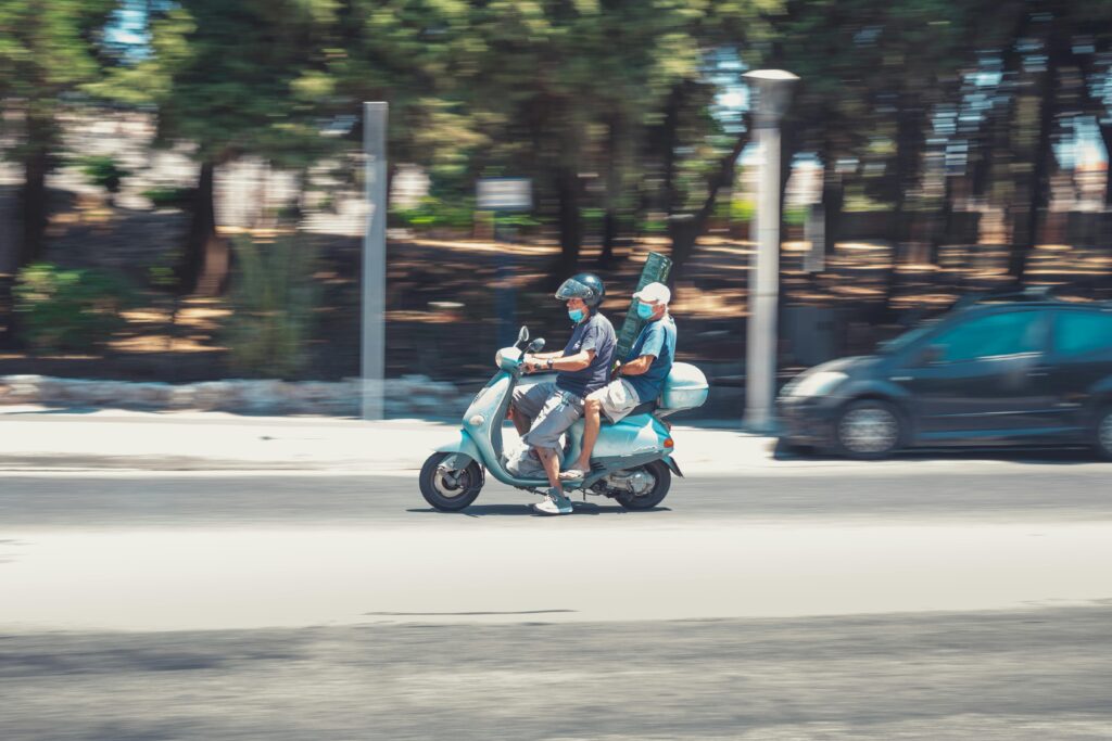 Two men wearing face masks ride a scooter on a sunny street in Greece, showcasing urban travel.