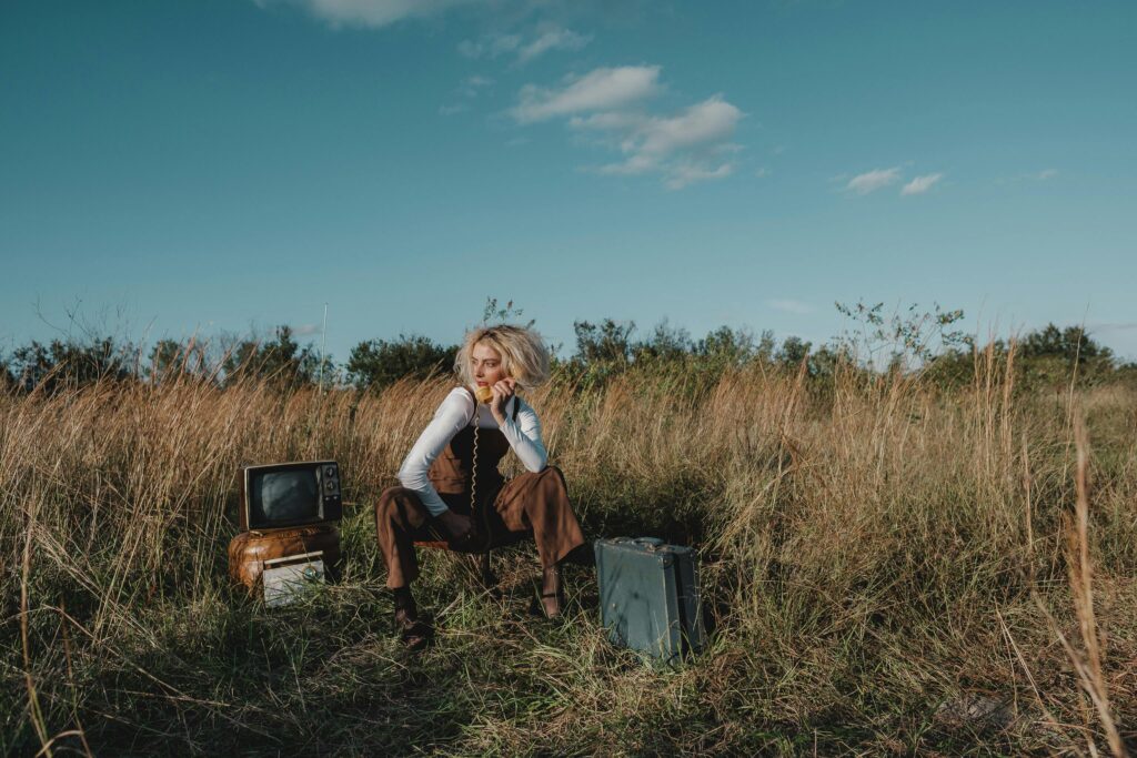 Stylish woman posing with vintage TV and suitcase in a sunny grassland setting.