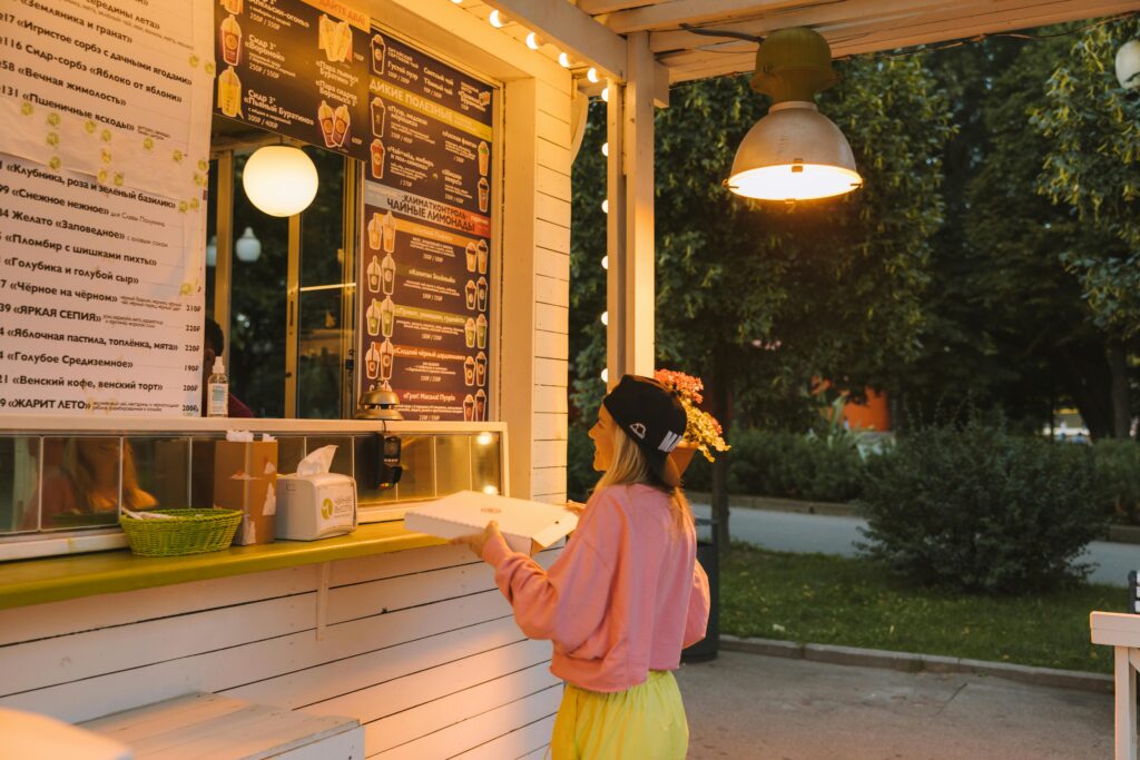 A young woman collects her pizza order at an outdoor food stall on a warm evening.