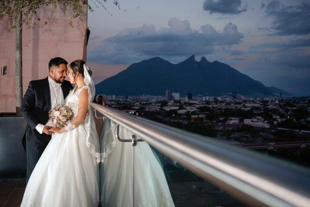 Bride and groom embrace lovingly on a balcony with stunning mountain backdrop at sunset.