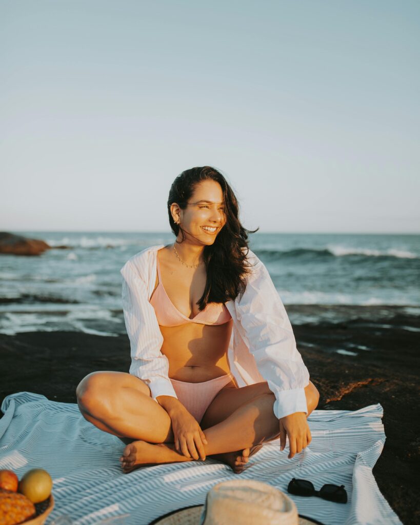 A woman in swimwear enjoys a sunny beach picnic by the sea, exuding relaxation and happiness.