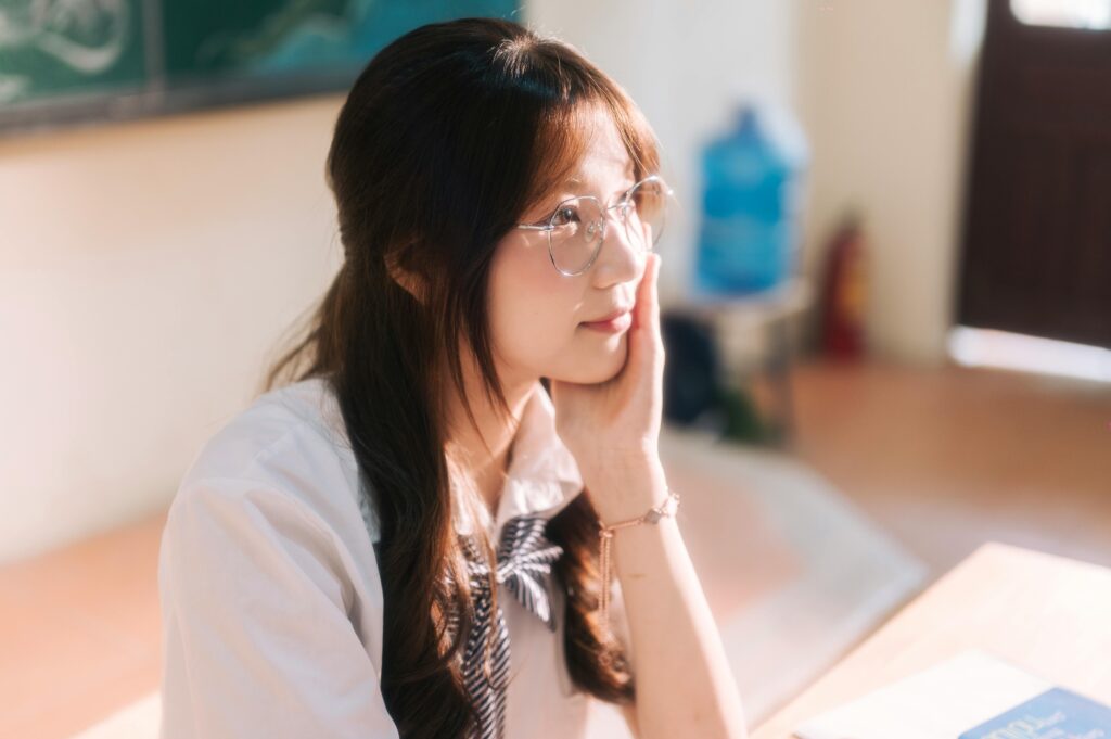 Young woman in glasses contemplating at desk in warm lit classroom setting.