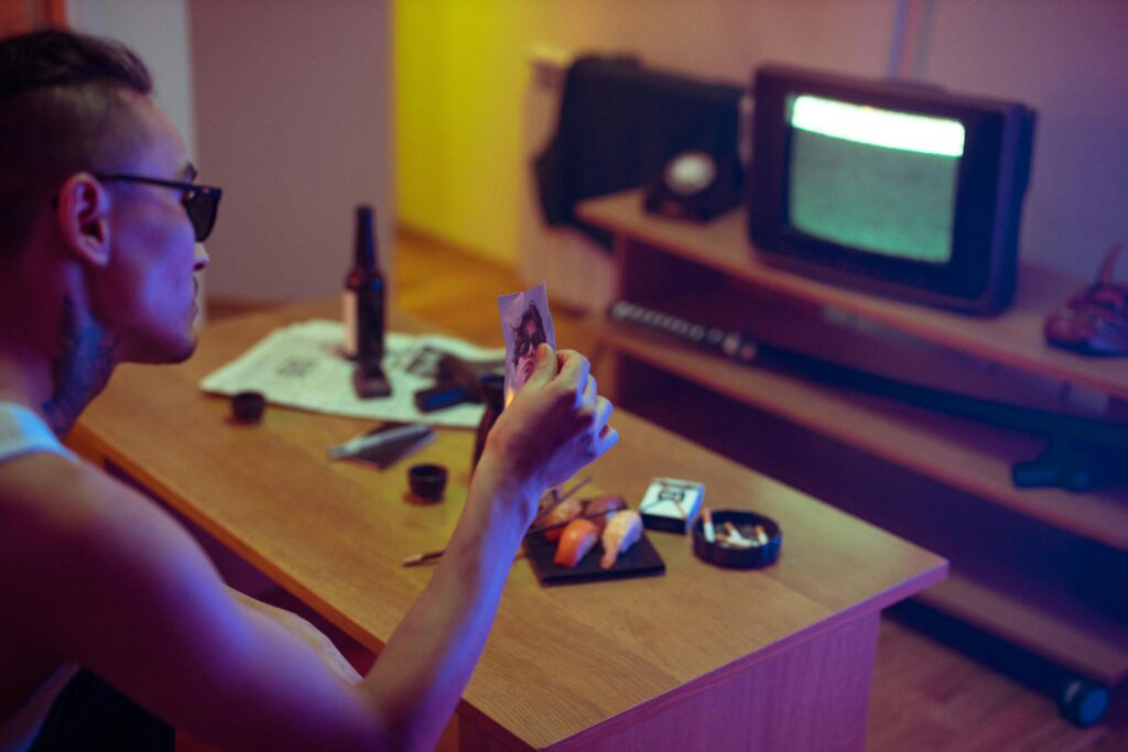 A tattooed man examines a photo in a dimly lit room with a retro TV.