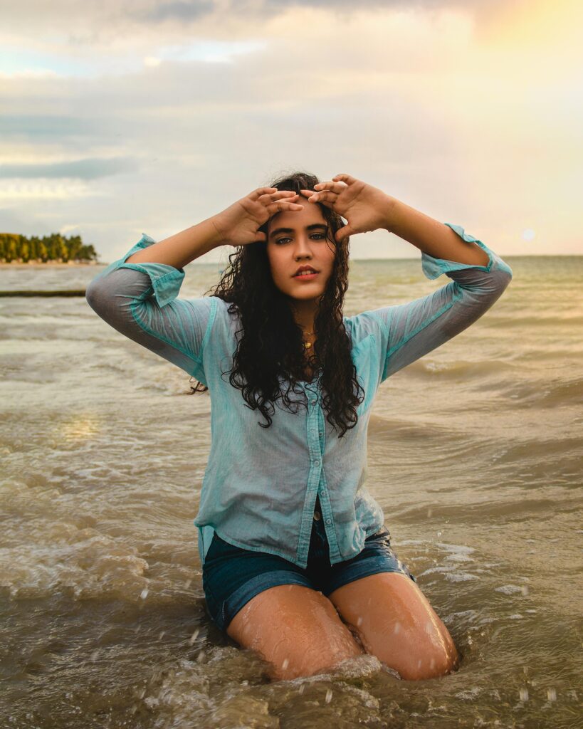 A woman with curly hair poses in the sea at sunset, capturing elegance and natural beauty.