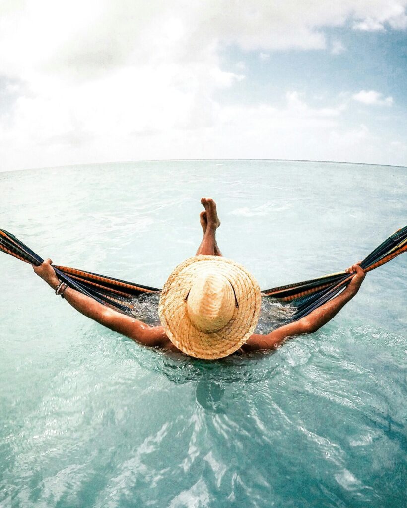 A person in a straw hat relaxes in a hammock above calm turquoise waters with a bright sky in the background.