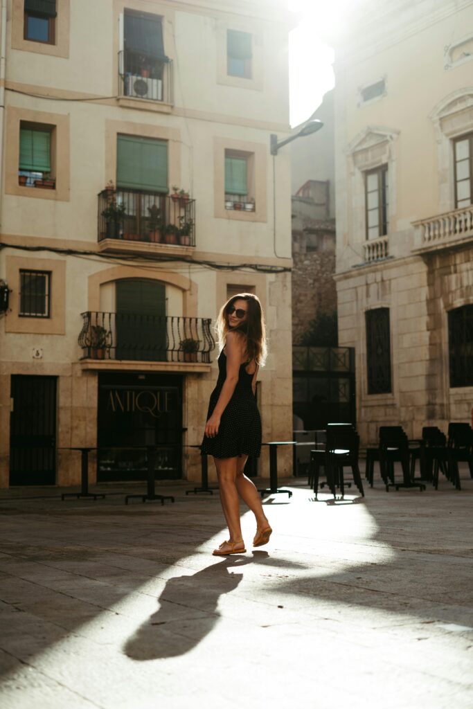 Stylish woman in sunglasses and black dress walks in sunlit Spanish street with chic architecture.