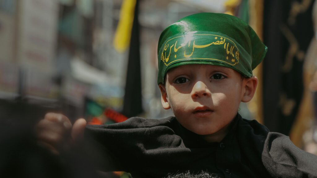 Portrait of a young boy in Srinagar wearing a green headband with Arabic script during a street event.