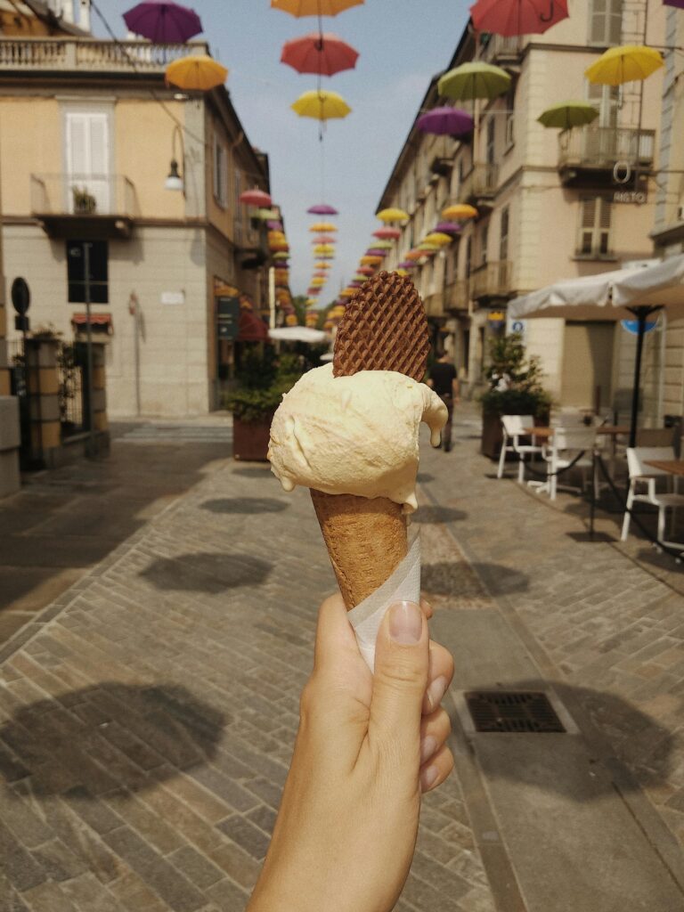 Hand holding vanilla ice cream cone with wafer in a vibrant umbrella-covered Italian street.