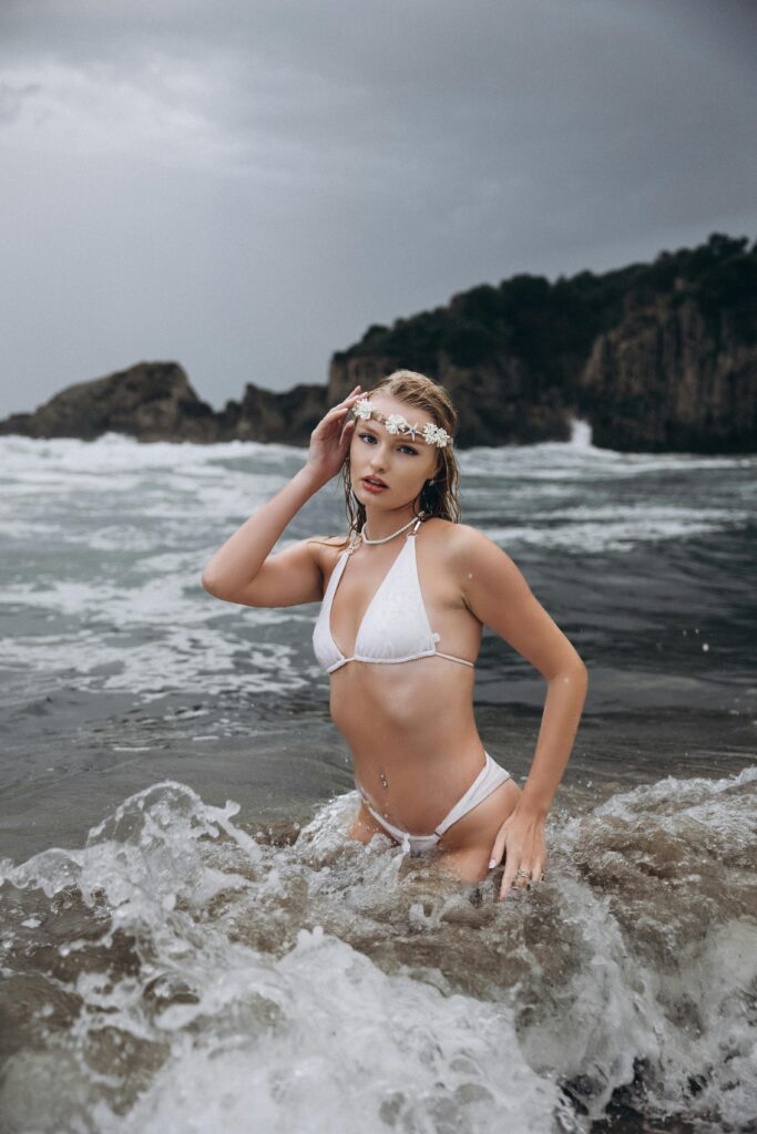 Woman in a white bikini posing in the ocean waves at a rocky beach.