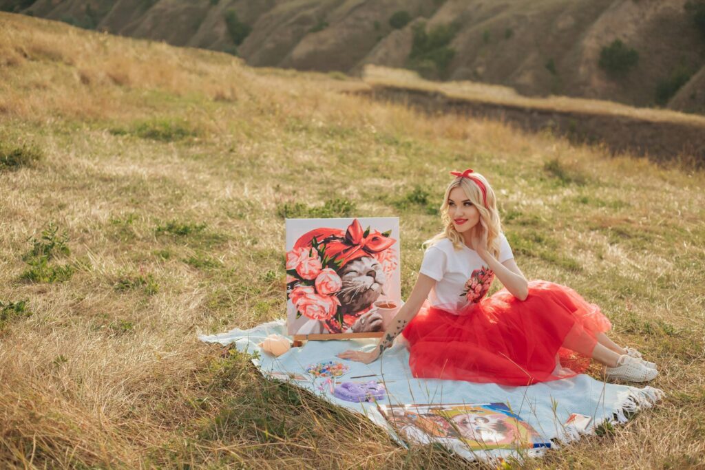 Blonde woman in a red tulle skirt painting outdoors in a scenic meadow.