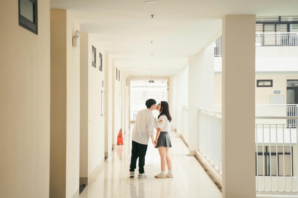 Asian couple shares a kiss in a brightly lit school corridor.