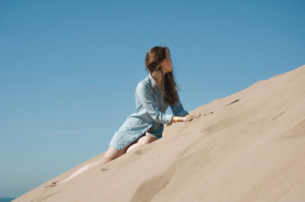 Woman in casual attire climbing sand dunes under a clear blue sky in Morocco.