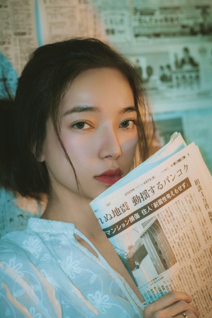 Close-up of a young woman reading a Japanese newspaper inside a room with newspaper-covered walls.