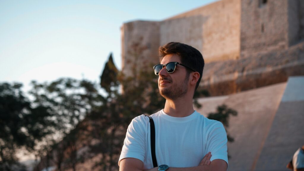 Man in sunglasses, white t-shirt enjoying sunset outdoors near a stone wall.