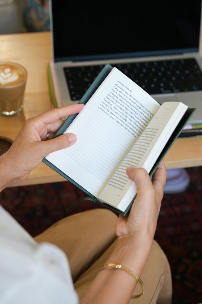 Person reads a book in a cozy cafe setting with an open laptop and coffee nearby.
