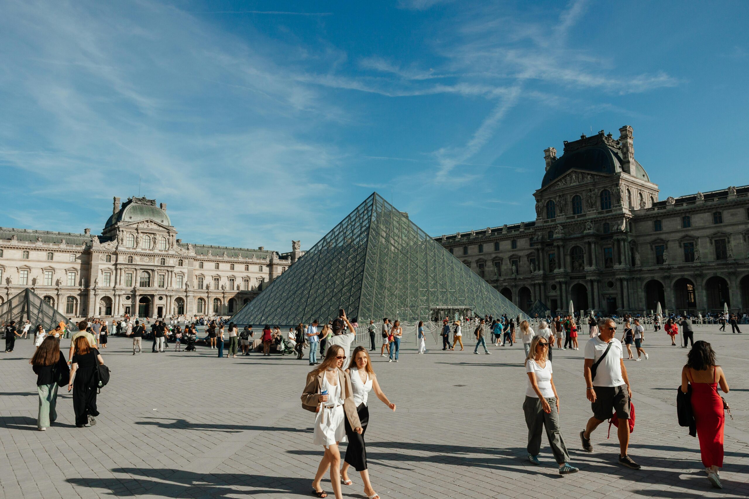 Crowds gather around the iconic glass Pyramid of the Louvre Museum in Paris, France on a sunny day.