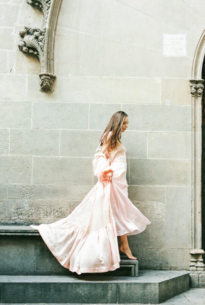 A woman gracefully walks barefoot in a flowing pink dress in a historic street setting.
