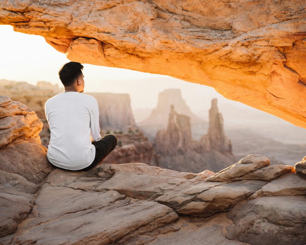 Man sitting under Mesa Arch admiring canyon view at sunrise, capturing the essence of adventure.
