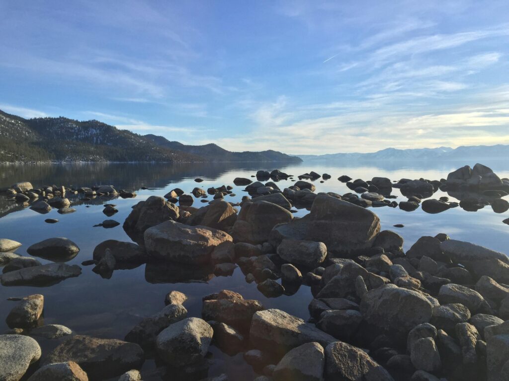 Tranquil mountain lake scene featuring calm water and scattered rocks under a clear blue sky.