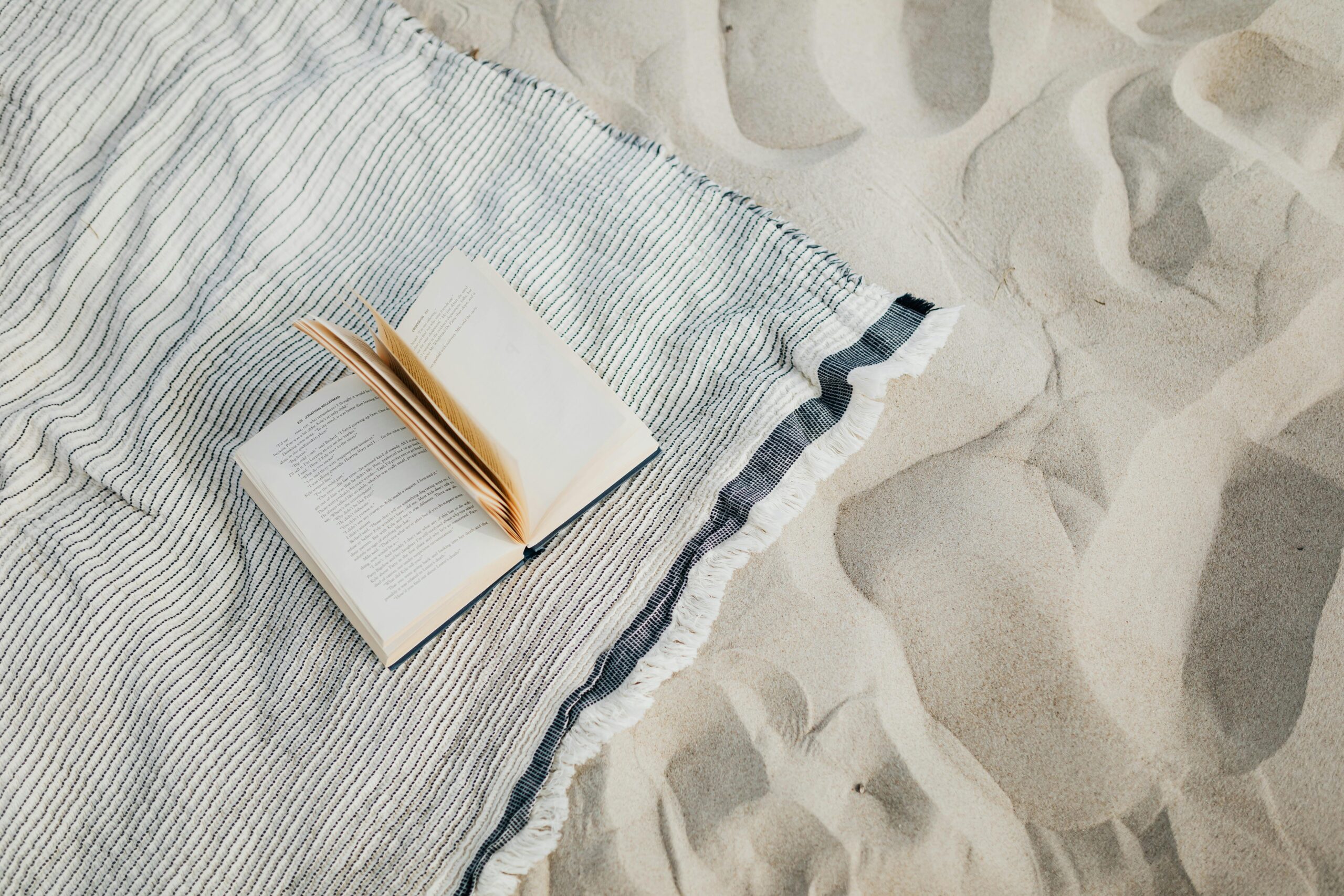 Open book resting on a striped beach towel on a sandy beach, calm setting.