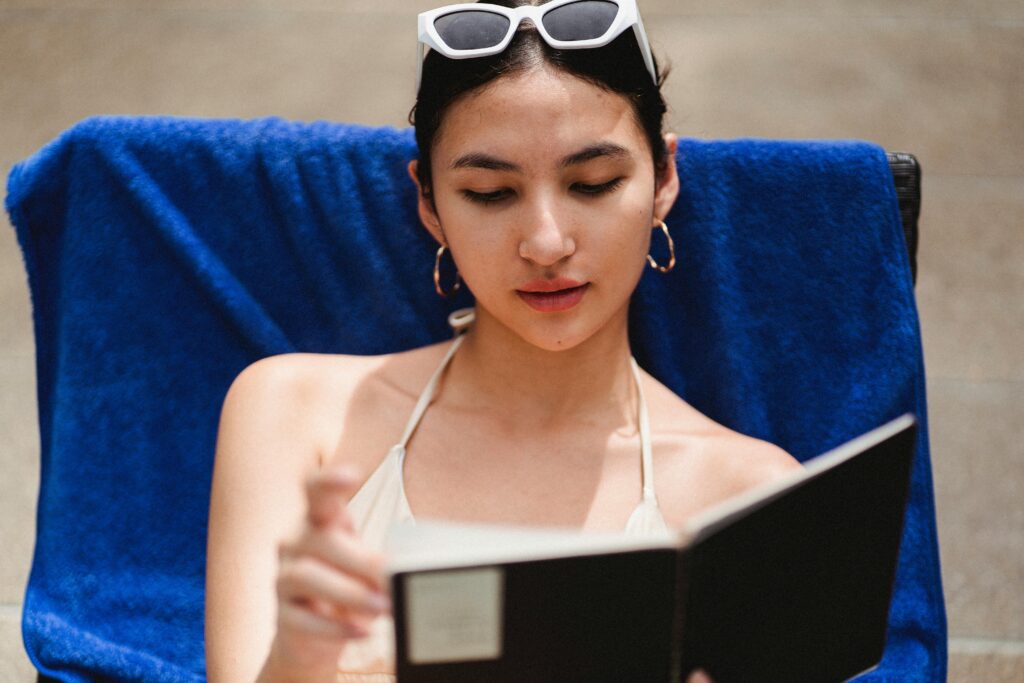 A young woman in swimwear reads a book by the pool, enjoying a sunny day outdoors.
