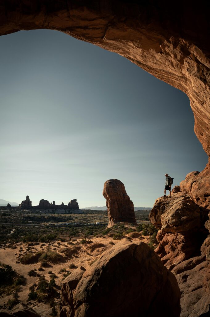 A lone hiker stands on a rock ledge, overlooking Arches National Park's stunning landscape.