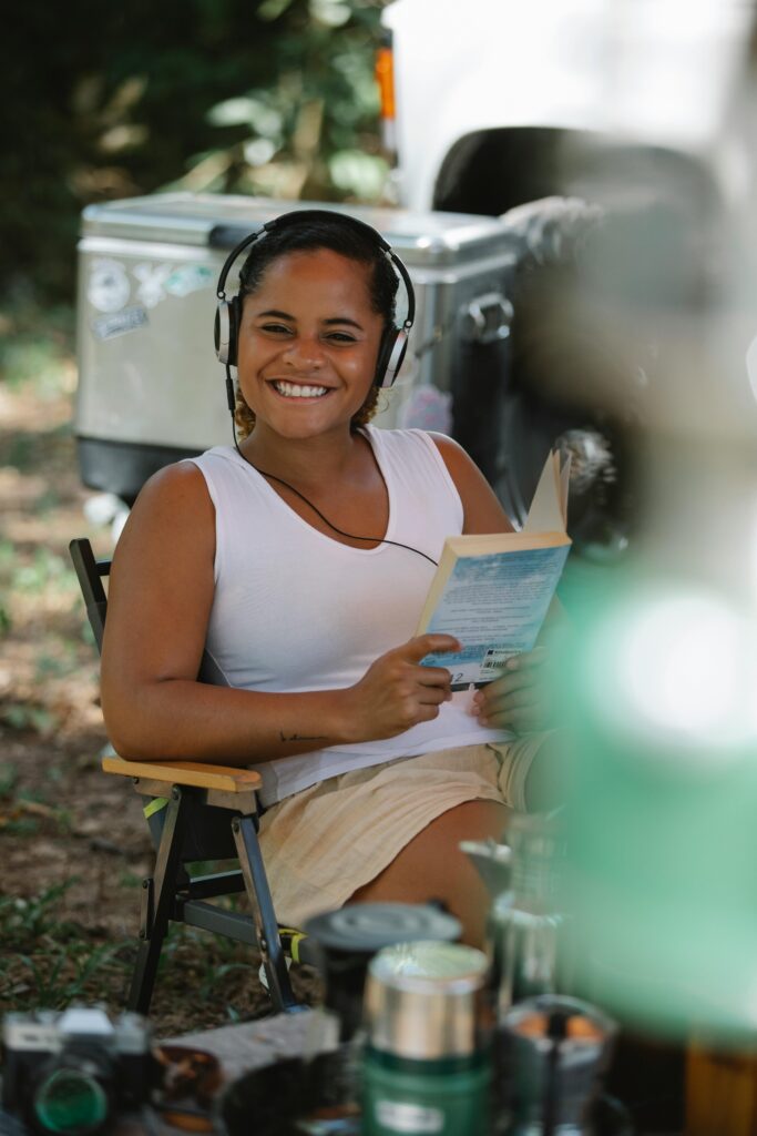 Joyful young tanned ethnic female tourist in casual clothes smiling and looking at camera while listening to music in headphones and reading novel at campsite on sunny day