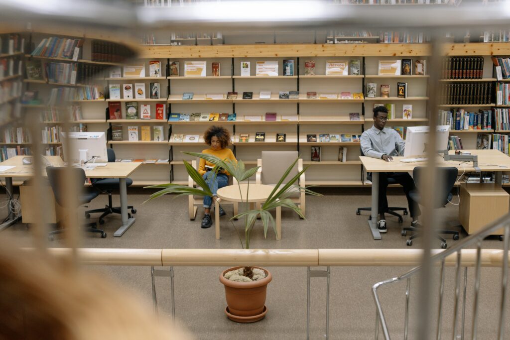 Indoor library scene with diverse students studying at desks surrounded by bookshelves.