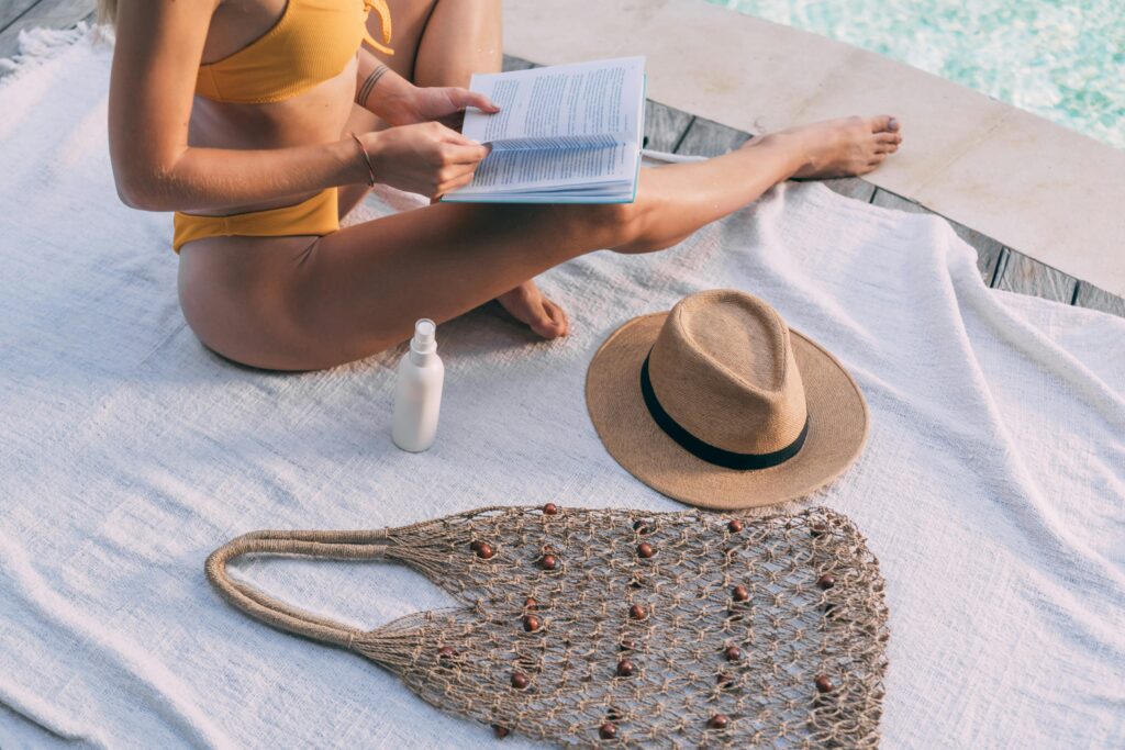 Adult female in swimwear relaxing by the pool, reading with a summer hat and bag nearby.