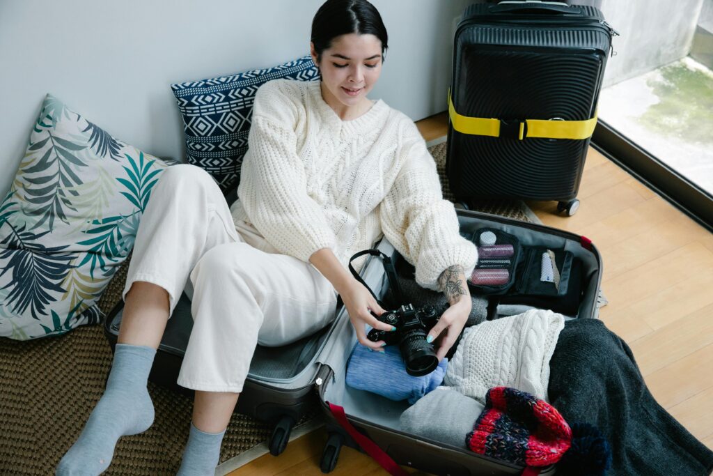 Young woman sitting on the floor packing a suitcase with clothes and a camera, preparing for a trip.