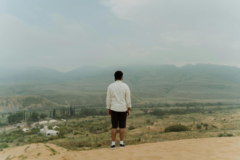 A man in casual attire stands on a desert edge, gazing at a serene mountainous landscape.