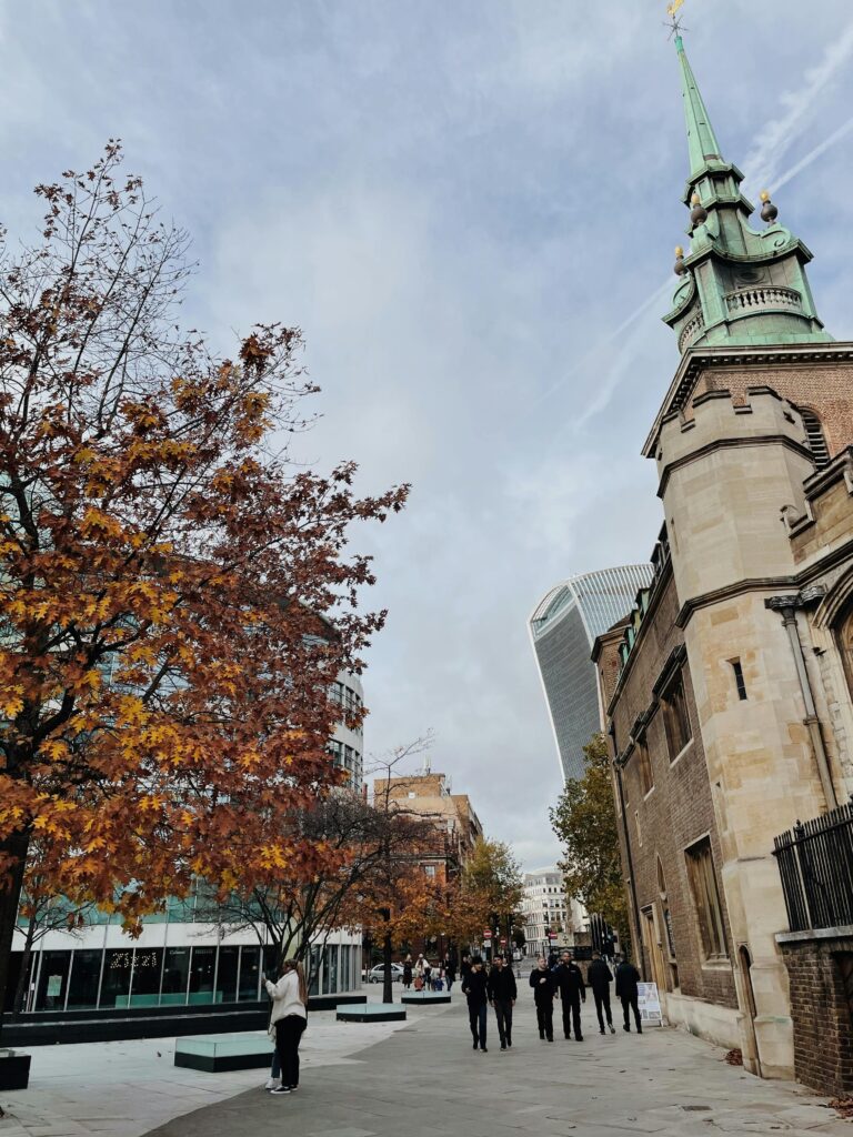 Urban scene with historic building, modern skyscraper, and autumnal trees under cloudy sky.