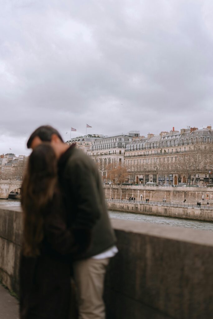 A couple shares a tender moment along the Seine River, with classic Parisian architecture in the backdrop.