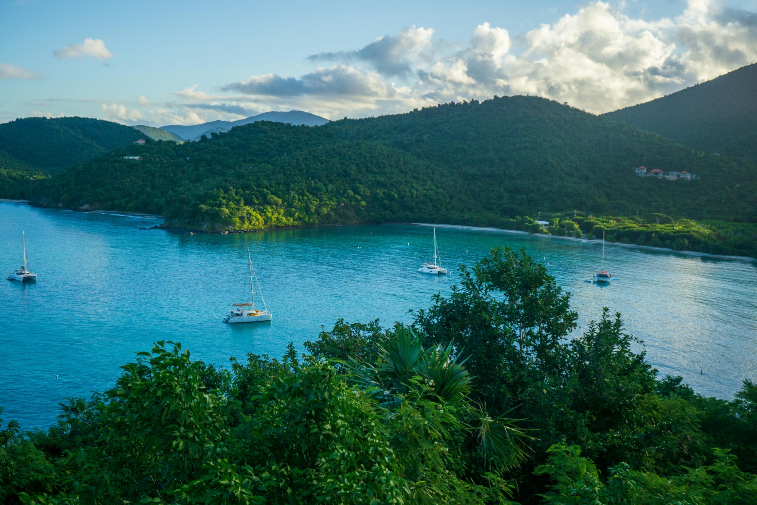A serene view of a tropical bay with sailboats, lush green mountains, and calm blue waters.