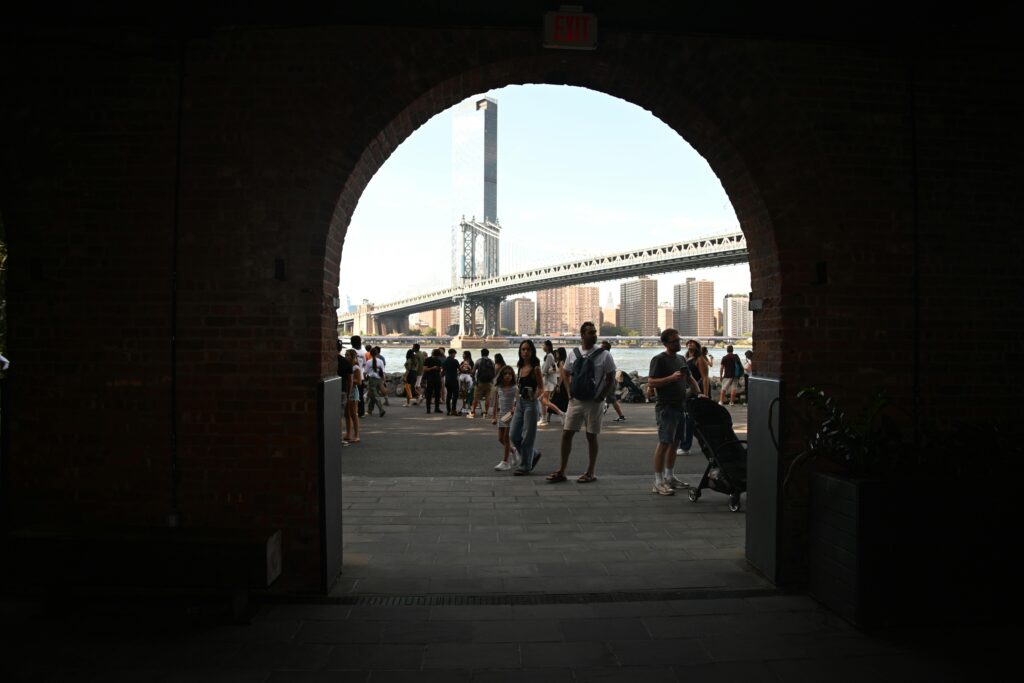 People gather near Manhattan Bridge on a sunny day, framed by a brick archway.
