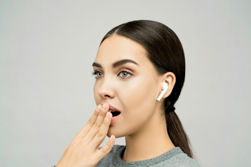 Portrait of a young woman with wireless earbuds, expressing surprise against a gray background.