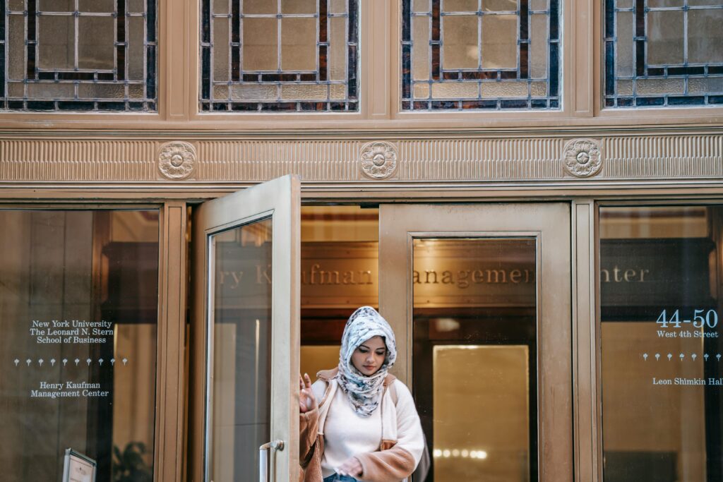 Ethnic young female in warm clothes and hijab walking out of management center door with glass walls in city street in daytime