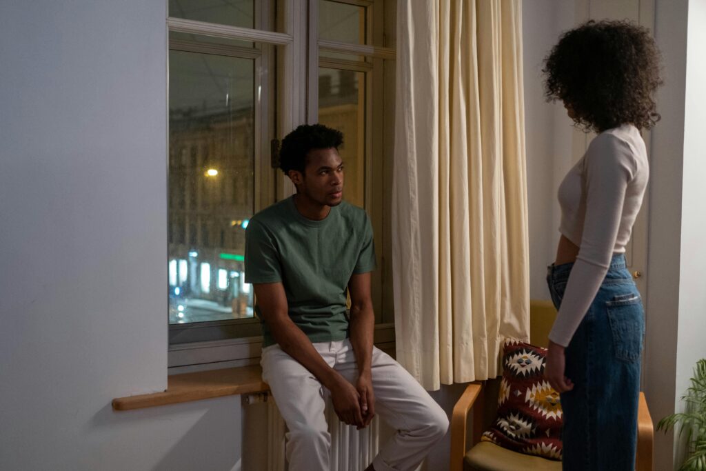 A couple engaged in a tense conversation by a windowsill in a cozy living room setting.