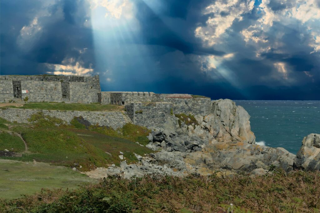 Rugged fort along Guernsey's rocky coast with dramatic clouds and sunrays breaking through.