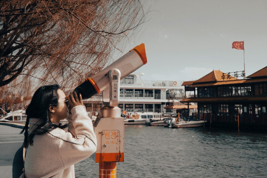 Asian woman looks through telescope by river with distant landmarks and boats.