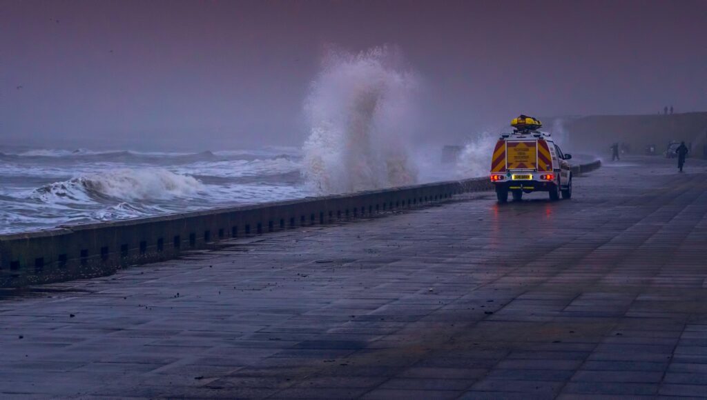 A dramatic coastal scene with waves crashing against a seawall and a vehicle on a stormy day.