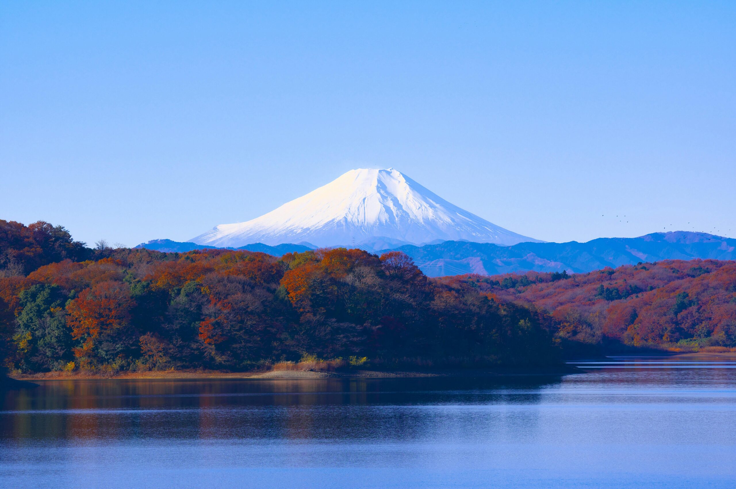 Breathtaking autumn view of Mount Fuji with calm lake and colorful trees reflecting clear sky.
