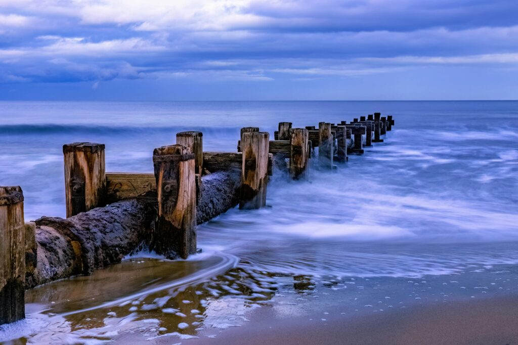 A tranquil seascape featuring a weathered wooden breakwater extending into the calm ocean under a cloudy sky.