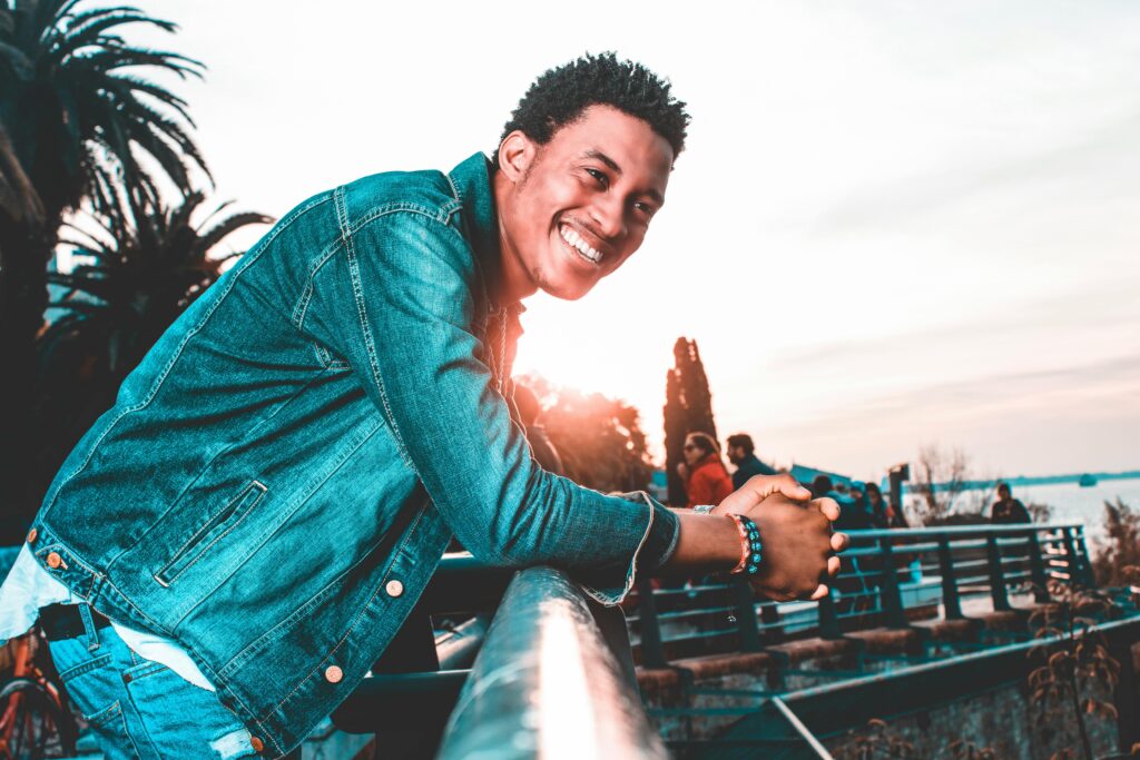 A man in a denim jacket leaning on a railing and smiling during a sunset outdoors.