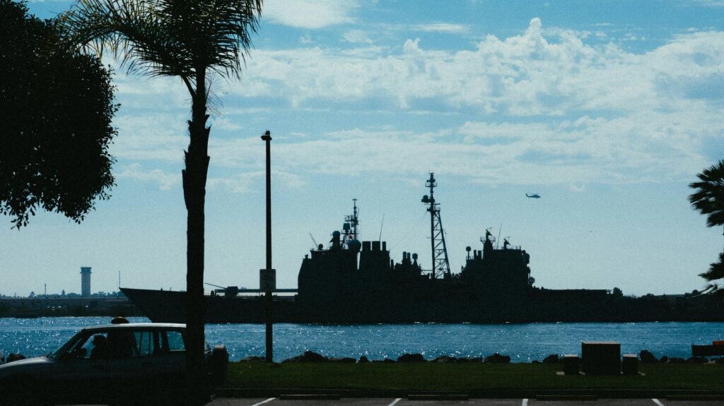 Silhouette of a navy warship in daylight, framed by palm trees and a clear sky.