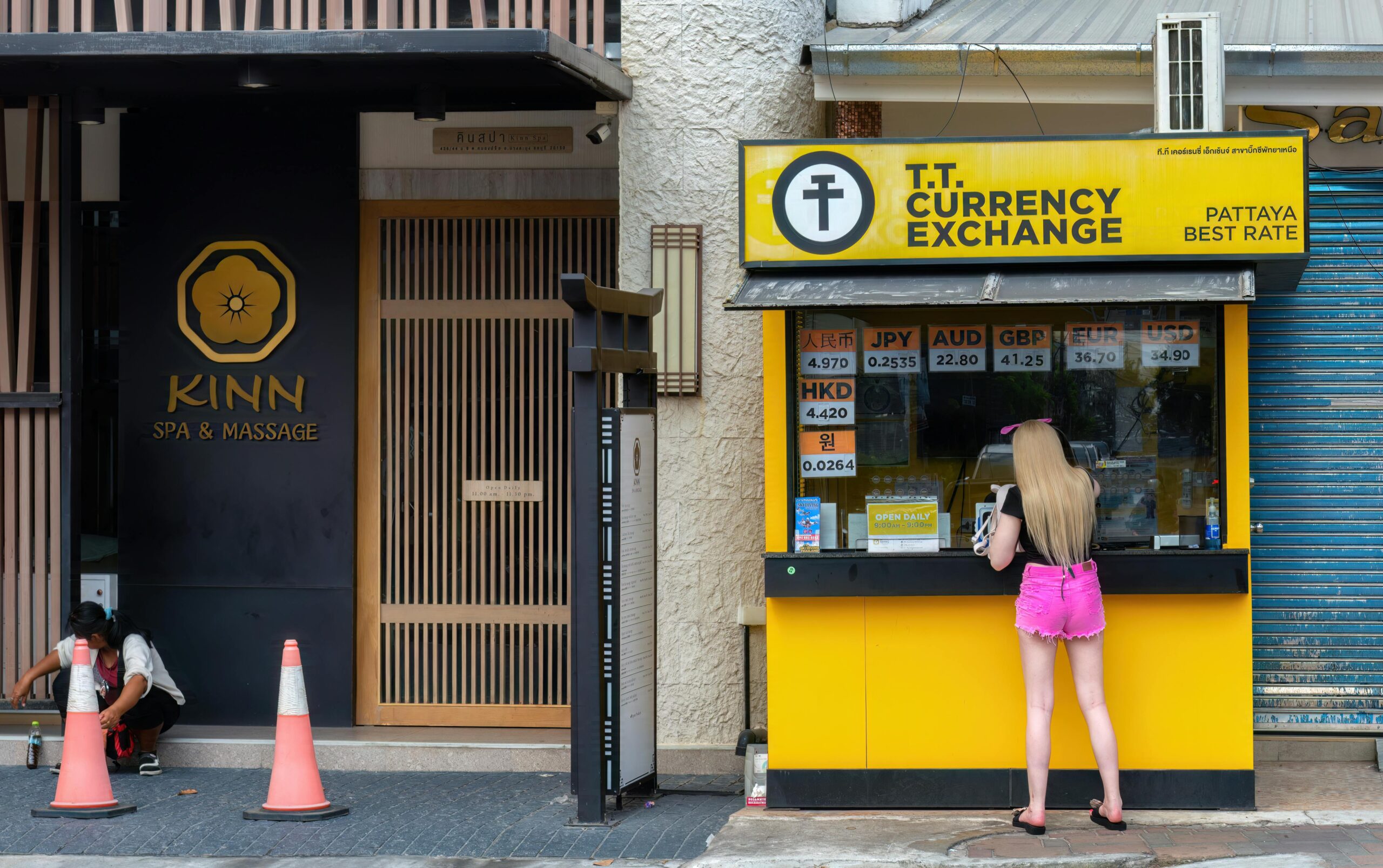 Woman at currency exchange booth in Pattaya, Thailand, with currency rates displayed.