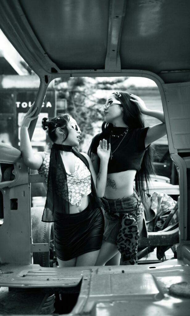 Two stylish women in trendy outfits pose inside a vehicle frame, expressing bold fashion choices.